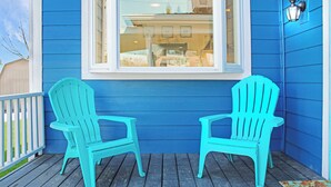 Two turquoise Adirondack chairs on a wooden deck with a blue exterior wall and a window above them.
