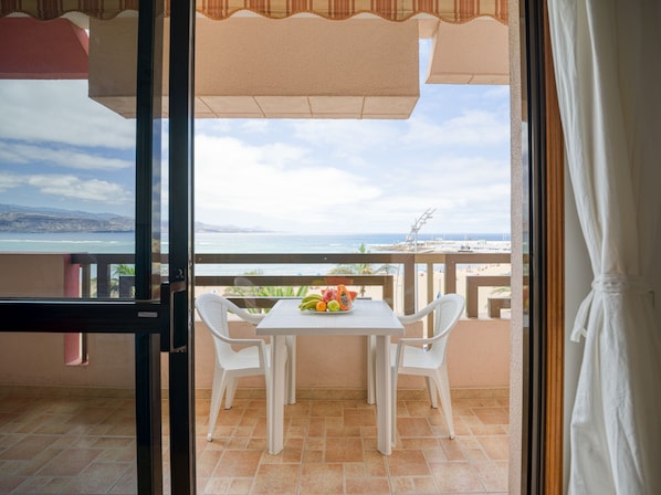 An open balcony with a table and chairs, a view of the sea, and a mountainous landscape in the distance.