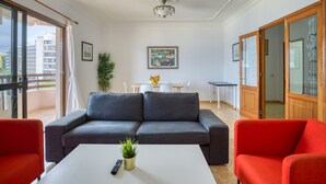 A living room with a grey sofa, red armchairs, a white coffee table, and a small plant. There is a dining area with a table and chairs in the background, and a balcony with a view of buildings. - Huge Balcony over Las Canteras By CanariasGetaway (Las Palmas de Gran Canaria)