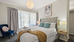 A hotel room with a large bed, a blue chair, a bedside table with a lamp, and a painting on the wall.