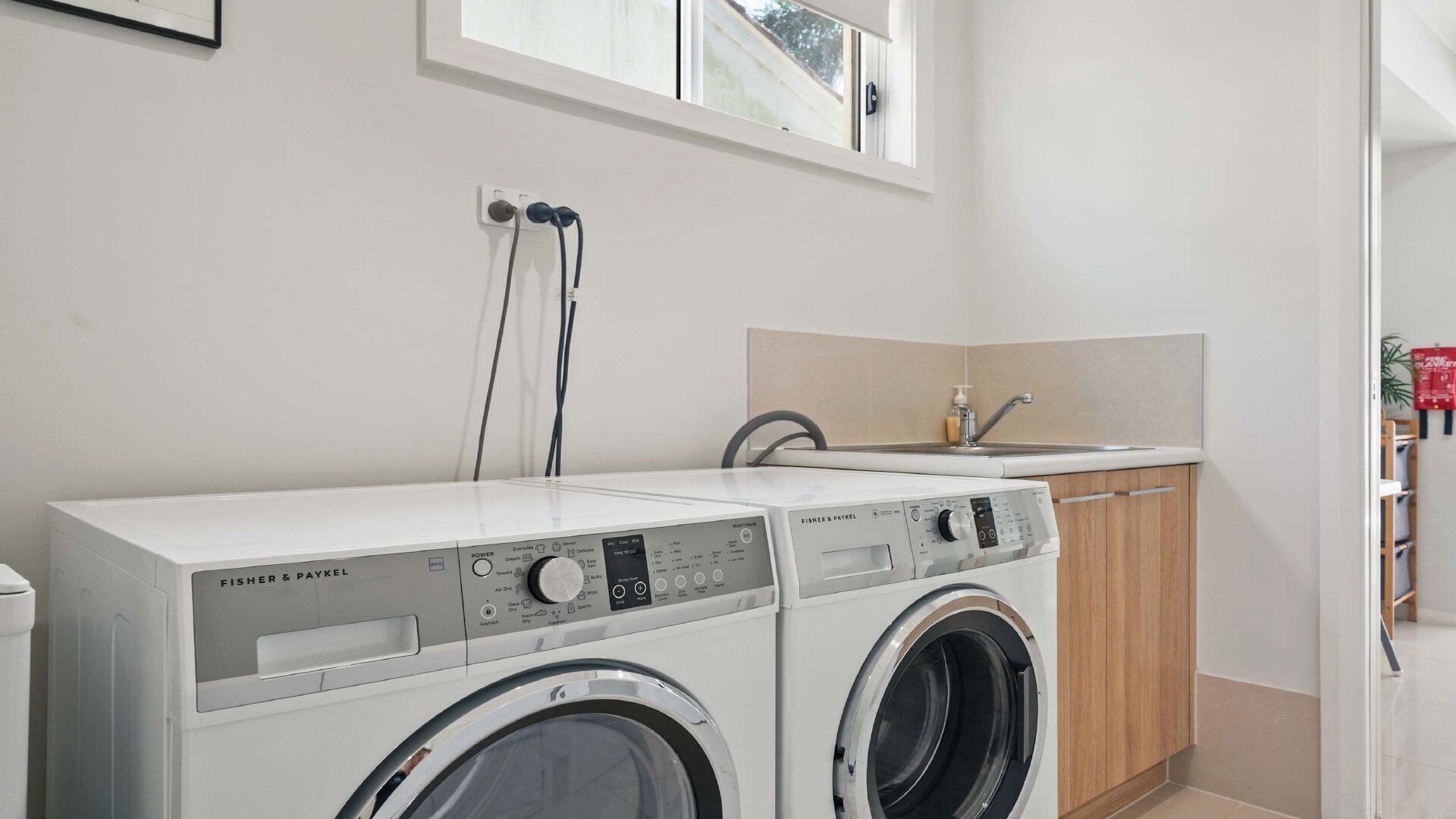 Un espace buanderie comprenant deux machines à laver blanches de la marque Fisher & Paykel, un meuble en bois, un évier et une fenêtre située au-dessus des machines.