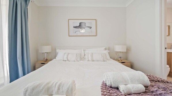 A bedroom with a large bed, two bedside tables with lamps, a framed picture on the wall, and a window with blue curtains. - Sea Shell - Brand New House - Direct Beach Access (Noraville)