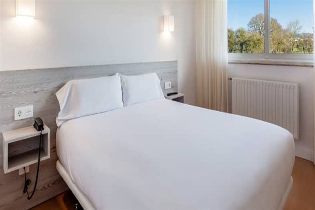 Una cama bien tendida en una habitación de hotel, con cabecera, mesitas de noche y ventana con vistas a árboles.