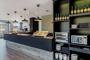 Una barra de desayuno moderna con diversas opciones de comida, una máquina de café y un enfriador de vino repleto de botellas.