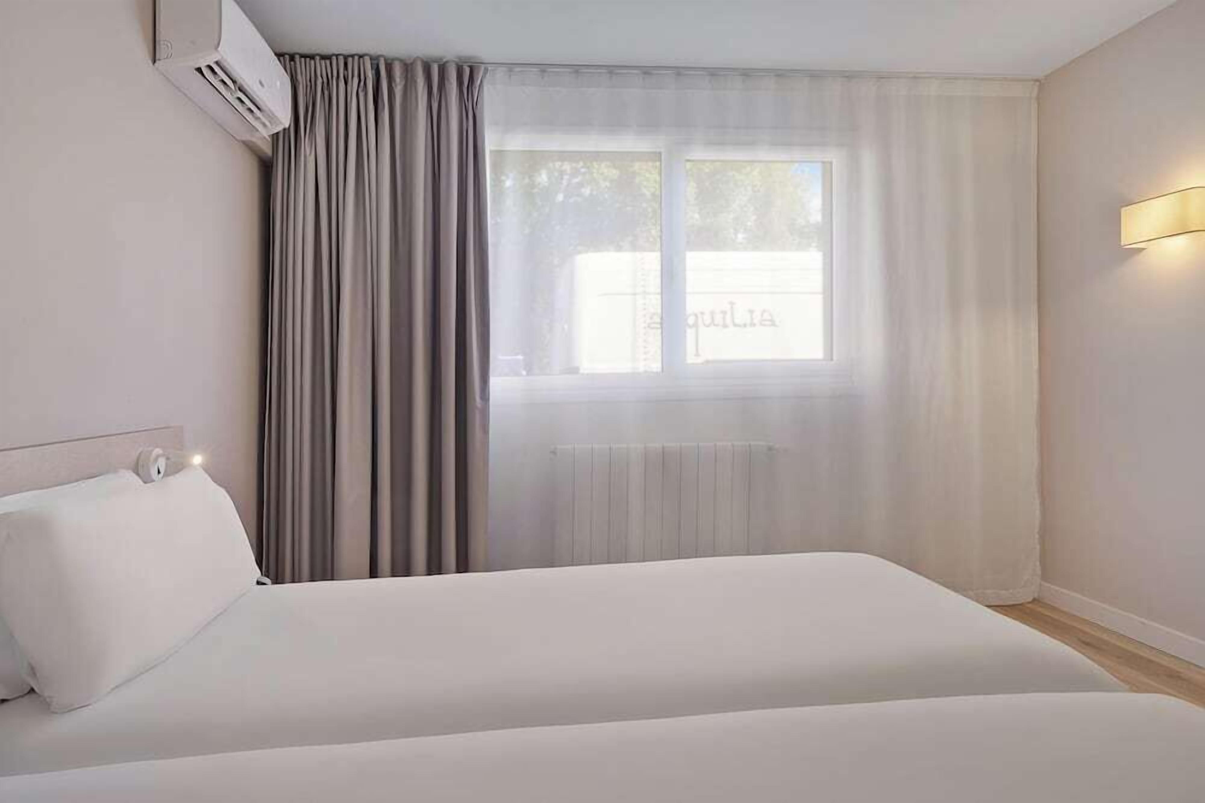 Habitación de hotel con cama, ventana con cortinas, equipo de aire acondicionado y lámpara fijada a la pared.