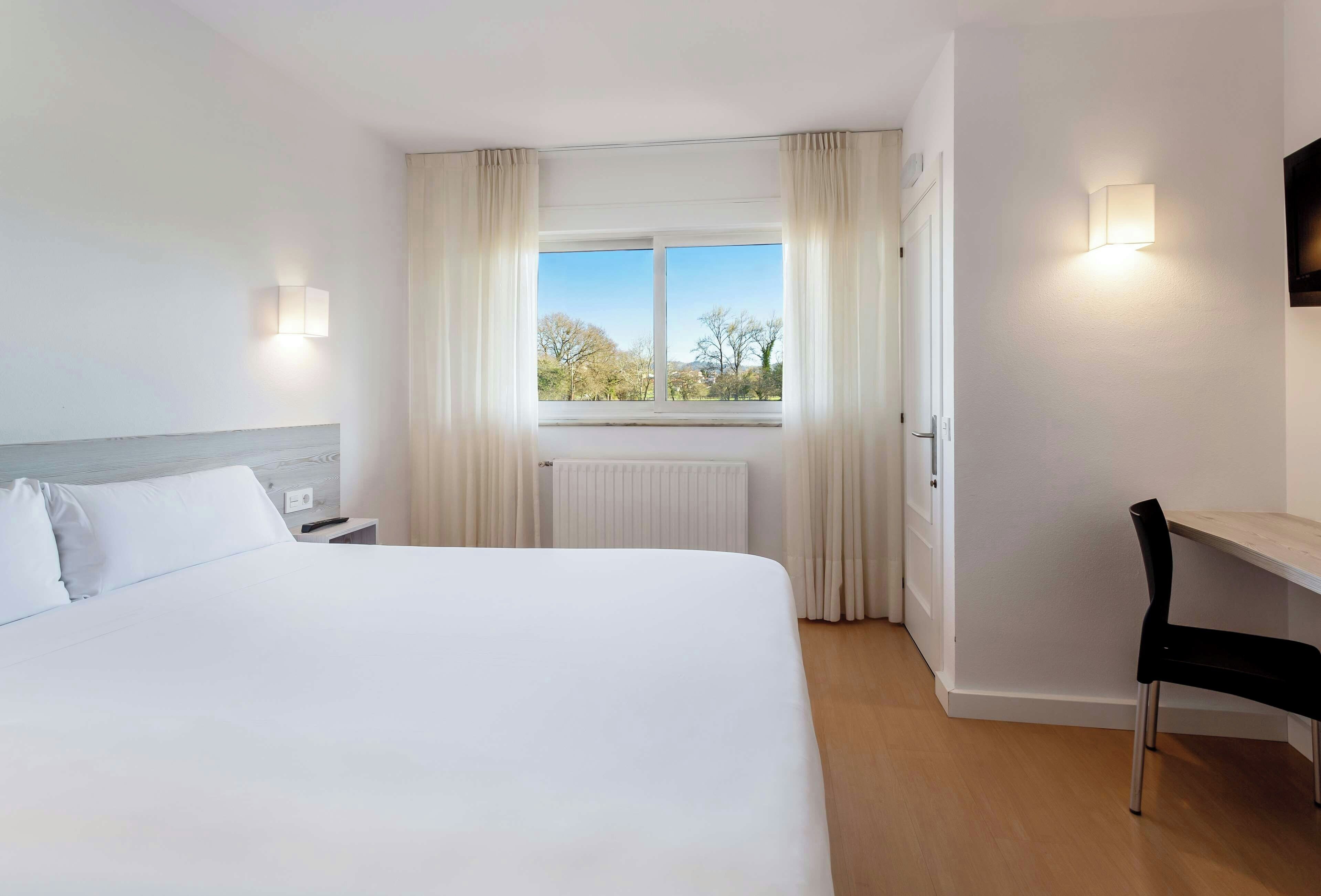 Une chambre d’hôtel avec un grand lit, un bureau, une chaise, une fenêtre avec des rideaux et un luminaire fixé au mur.