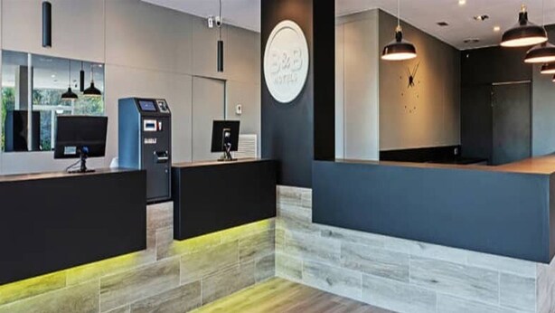 A modern hotel reception area with a reception desk, a digital check-in kiosk, and a clock on the wall. - B&B Hotel Oviedo (Siero)