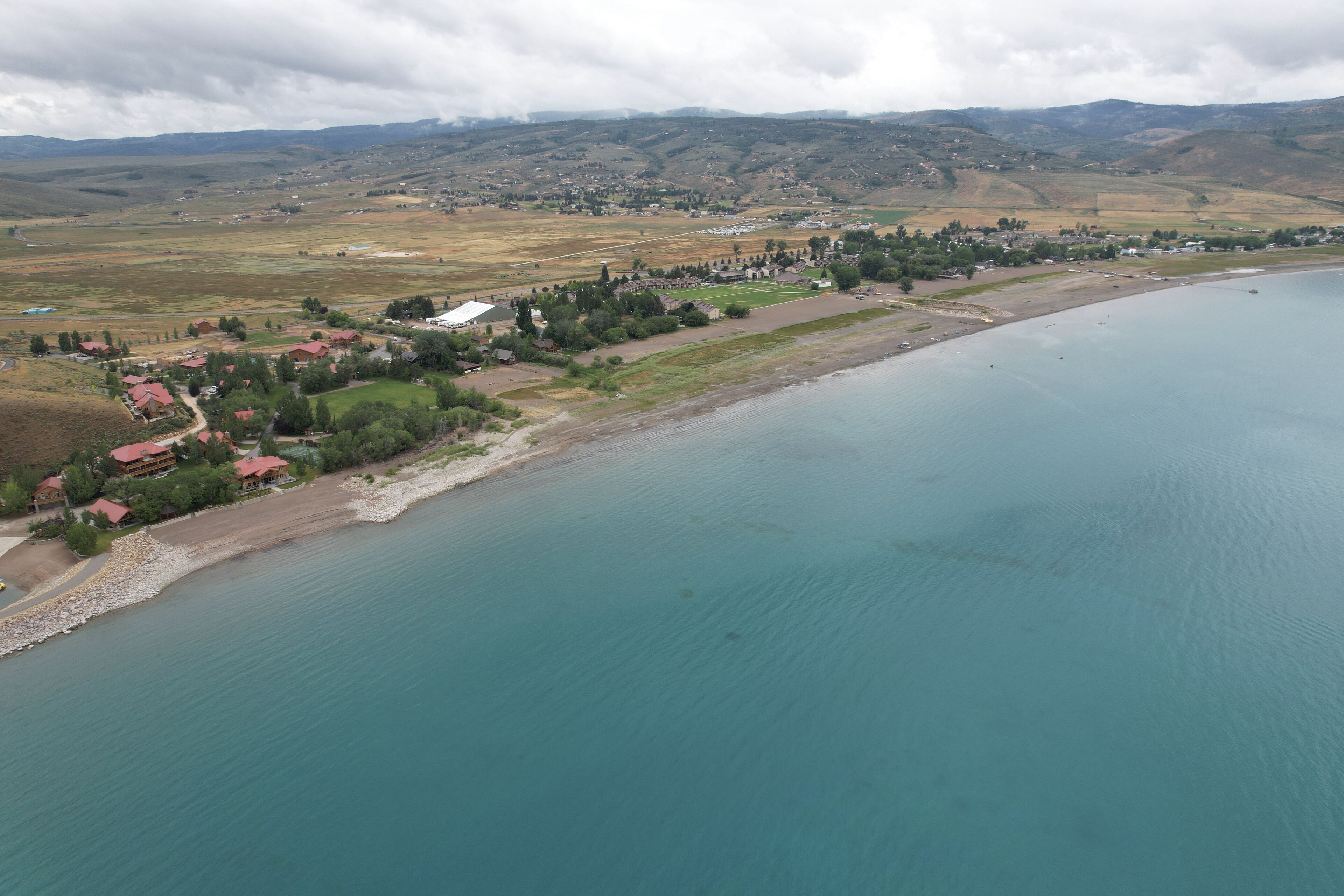 Vista aérea de un pueblo a orillas de un lago, con casas a lo largo de la costa, un pequeño pueblo al fondo y colinas circundantes.