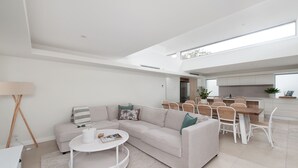 A modern living room with a sectional sofa, a round coffee table, and a dining area with white chairs and a wooden table. - Baydream on the Beach - With Breathtaking Water Views (Corlette)