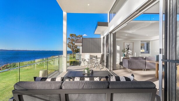 A modern balcony with a view of the sea, a dining area with a table and chairs, and a living area with a sofa and a coffee table. - Baydream on the Beach - With Breathtaking Water Views (Corlette)
