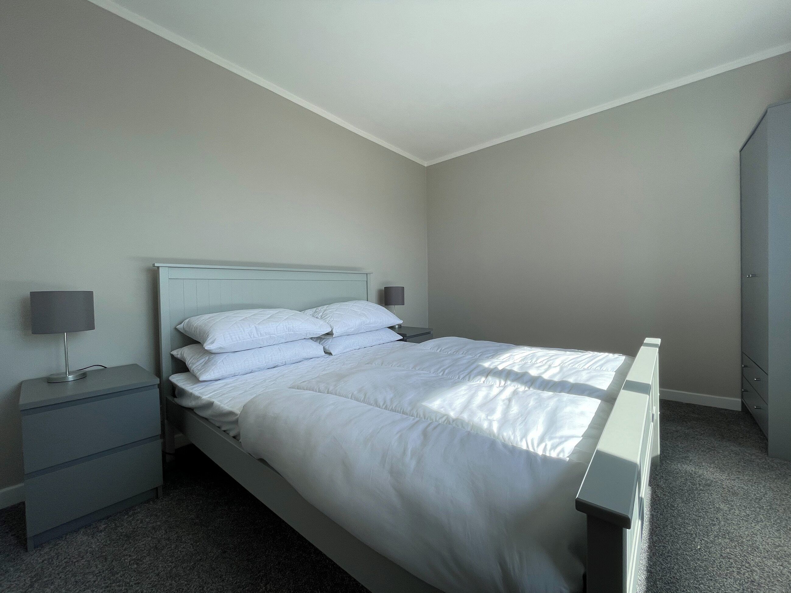 A neatly made bed with white bedding in a room with light-colored walls and a gray carpet. There is a nightstand with a lamp and a small drawer.