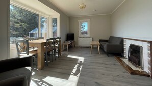 Un vaste espace de vie comprenant une table à manger, des chaises, un canapé, une cheminée et de grandes fenêtres donnant sur l’extérieur.
