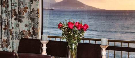 A dining table with a vase of red roses, two candles, and a view of the ocean and a mountain.