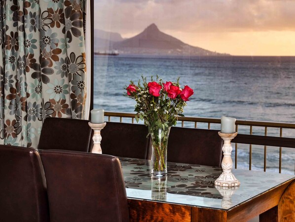 A dining table with a vase of red roses, two candles, and a view of the ocean and a mountain.