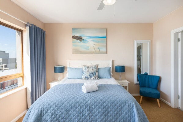 A bedroom with a large bed, two bedside tables, a chair, a painting, and a view of the outside.