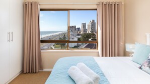 A hotel room with a large window, a bed with a blue quilt, and a view of a cityscape and beach. - Witsand 401 by CTHA (Cape Town)