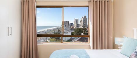 A hotel room with a large window, a bed with a blue quilt, and a view of a cityscape and beach.