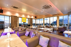 A modern hotel lobby with leather seating, a bar area, and large windows offering city views.