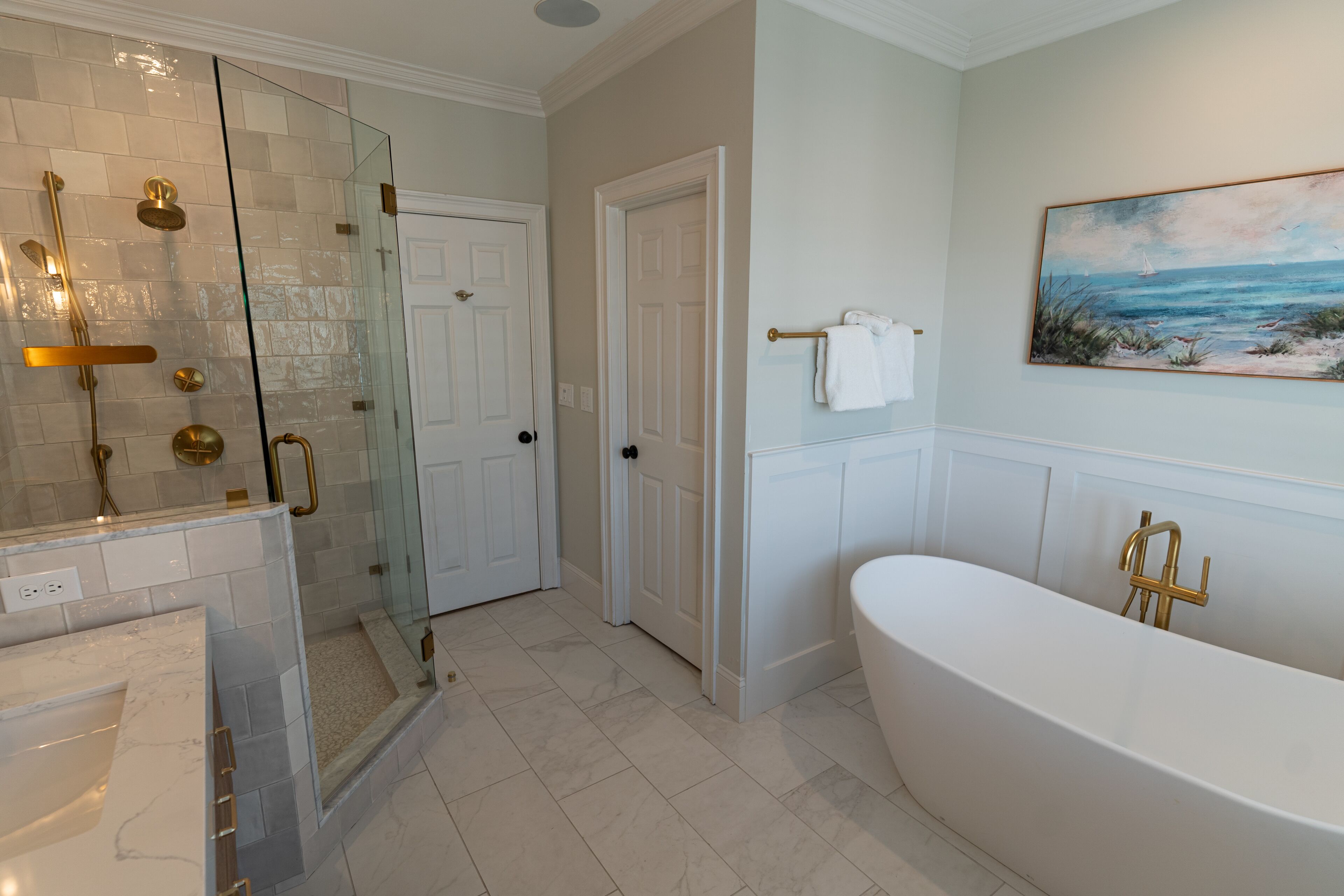 A bathroom with a glass-enclosed shower, a freestanding bathtub, and a painting of a beach scene on the wall.