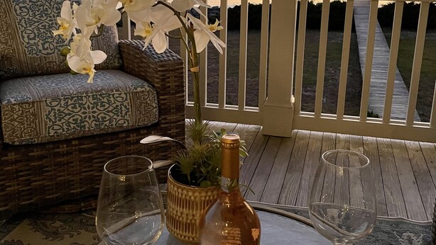 Une terrasse dotée d’un canapé en osier et d’une table sur laquelle sont disposés une bouteille et deux verres à vin, offrant une vue sur le coucher de soleil sur l’océan.