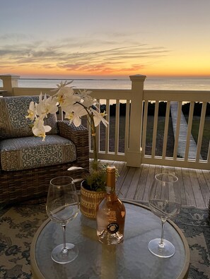 A deck with a wicker sofa and a table set with a bottle and two wine glasses, overlooking a sunset over the ocean. - Experience Seabrook Island bliss, Expansive ocean views, Dolphin lovers oasis (Seabrook Island)