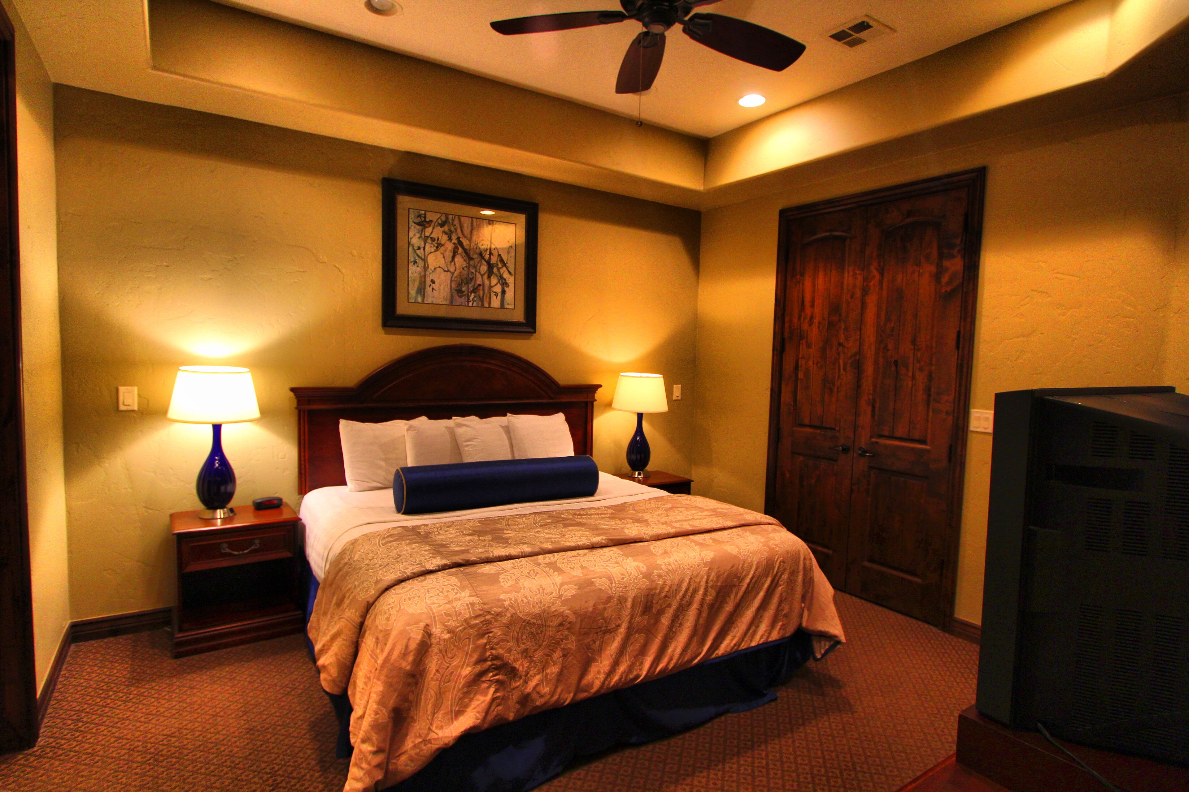 A hotel room with a large bed, two bedside lamps, a ceiling fan, a wooden door, and a television.