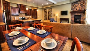 A dining area with a wooden table set for four, a kitchen in the background, a stone fireplace, and a ceiling fan. - Canyon Villas at Coral Ridge (Washington)
