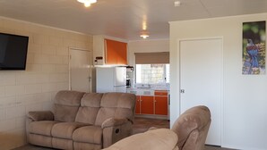Un salon avec deux fauteuils inclinables de couleur beige, un téléviseur à écran plat fixé au mur, et un coin cuisine avec des meubles orange et un réfrigérateur blanc.
