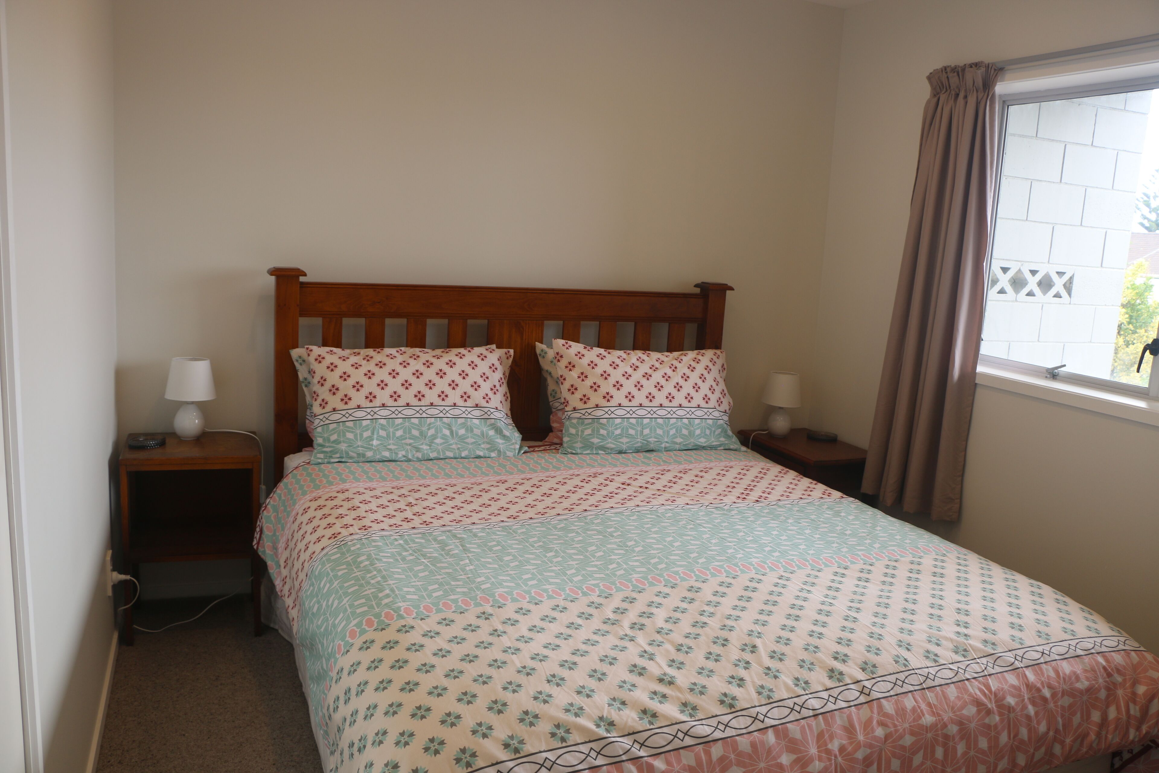 Une chambre à coucher avec un lit en bois, une literie à motifs floraux, une table de chevet avec une lampe et une fenêtre avec des rideaux.