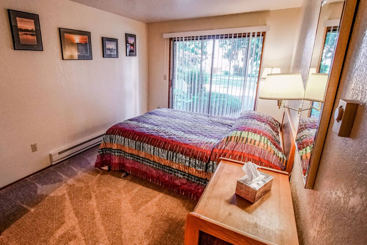 A bedroom with a large bed, a bedside table with a tissue box, and a window with blinds.