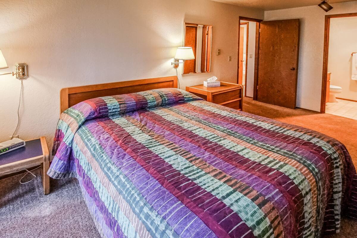 A hotel room with a large bed, a bedside table with a lamp, a nightstand, and a wall-mounted mirror.
