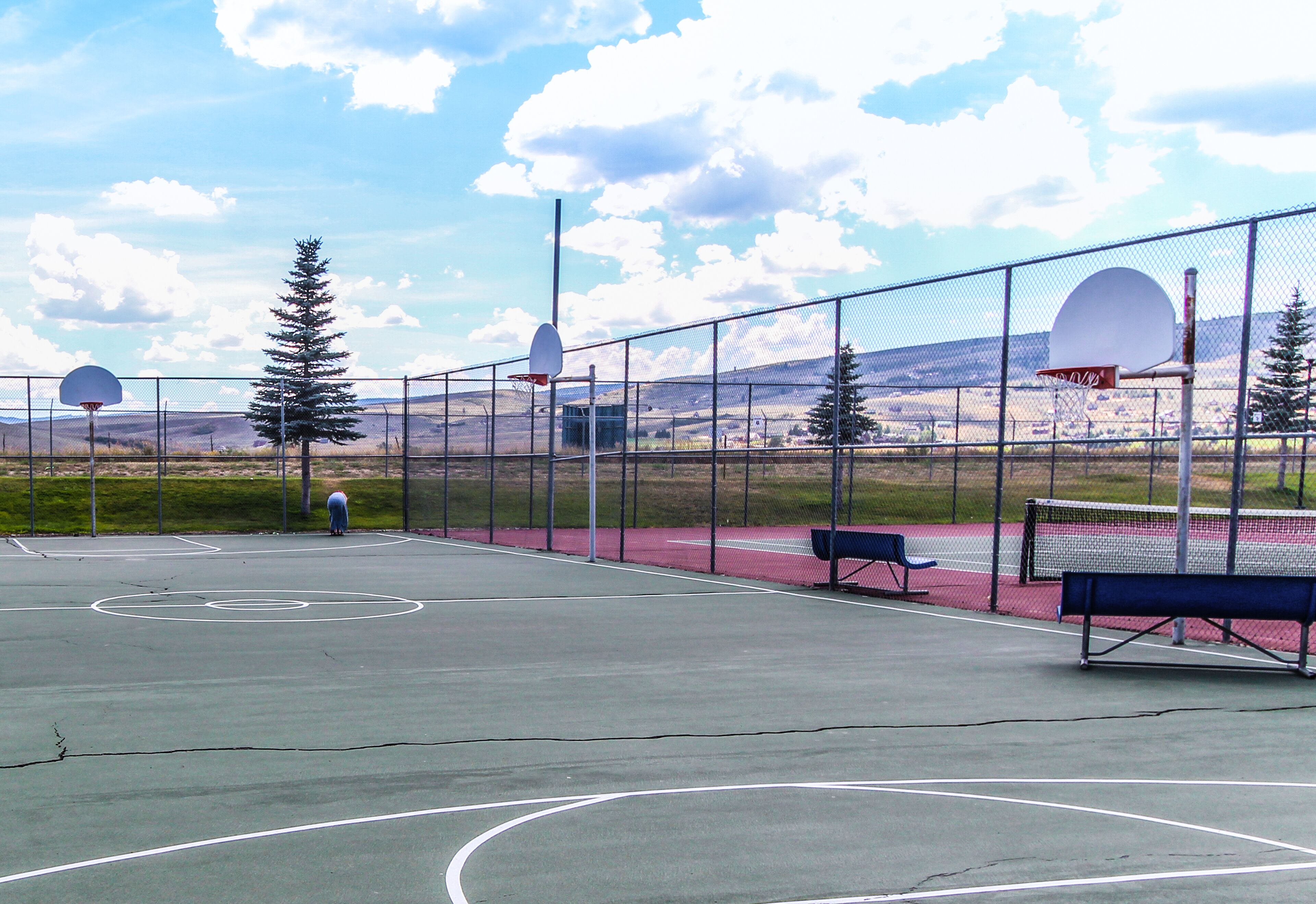 Quadra de basquete externa com cerca de arame, aros de basquete e bancos.