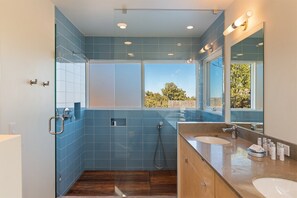 Ein modernes Badezimmer mit blauen Fliesen, einer Duschkabine aus Glas, Holzboden und einem großen Fenster mit Blick ins Freie.