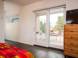 Ein modernes Schlafzimmer mit einem großen Bett, einer Holzkommode, einer Glastür mit Schiebeöffnung, die auf einen Balkon mit Sitzgelegenheit führt, und einem Fernseher auf einer Kommode.