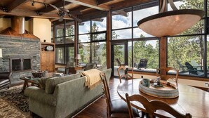 Un vaste salon doté d’une cheminée en pierre, de grandes fenêtres offrant une vue sur l’extérieur et d’un coin repas avec des meubles en bois.