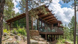 Une cabane dotée d’une grande fenêtre en verre, d’une terrasse en bois et de fondations en pierre.