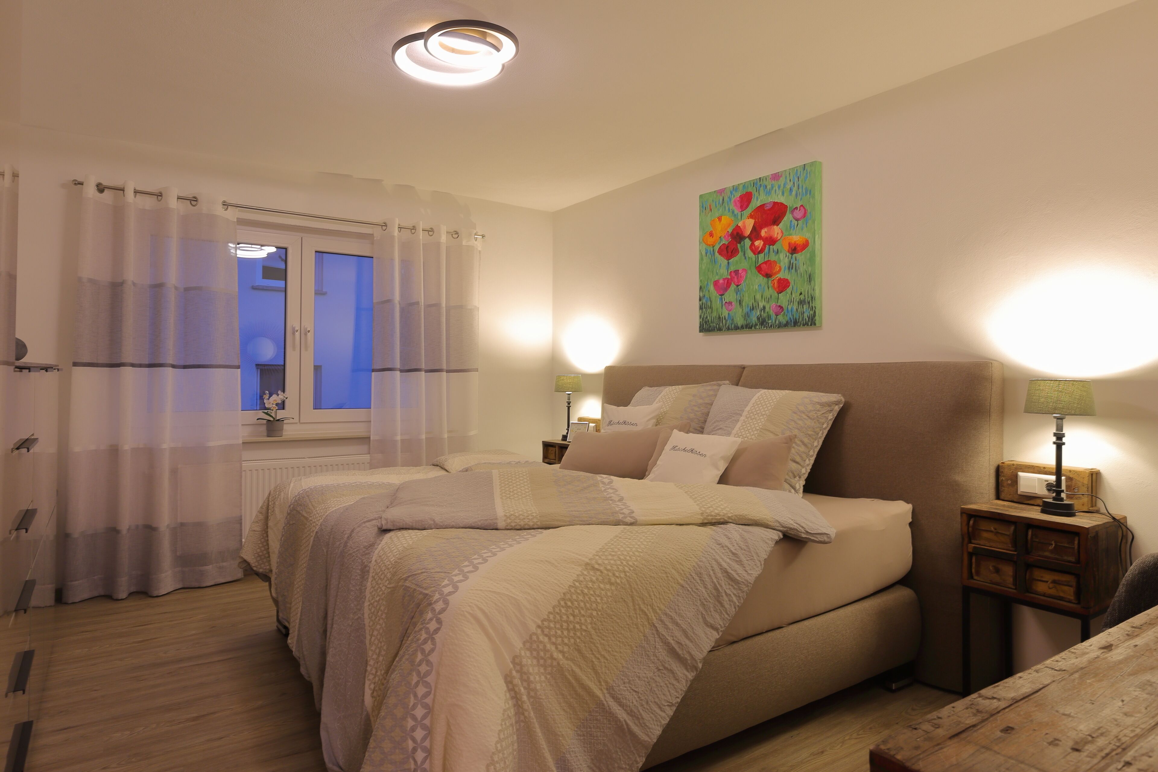 A bedroom with two beds, a window with curtains, a painting on the wall, and bedside lamps.