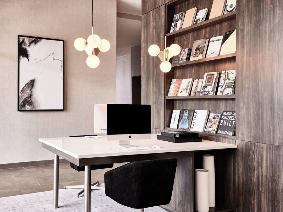 A modern office space with a white desk, a computer, a black chair, a bookshelf with books and magazines, and a framed abstract painting on the wall.
