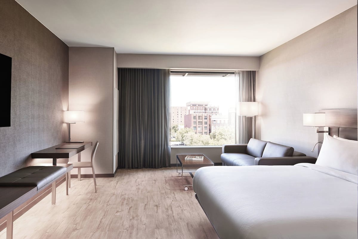 Una habitación de hotel moderna con una cama grande, un escritorio, una silla, un sofá y vistas a la ciudad desde la ventana.