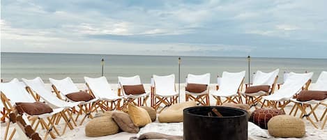 A beach with rows of wooden lounge chairs and a black grill in the sand.