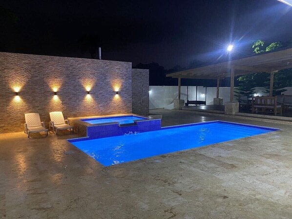 A swimming pool with a blue tint, surrounded by a concrete deck and a stone wall. There are two lounge chairs and a small seating area with a table.