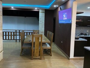Ein Essbereich mit Holztisch und -stühlen, eine Küchenzeile mit Arbeitsplatte und Spüle sowie ein an der Wand befestigter Fernseher.