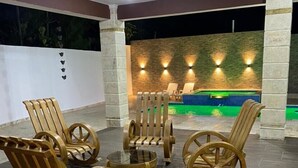 Area piscina con poltroncine a dondolo in legno e un piccolo tavolo, parete in pietra con luci a parete e piscina con bordo verde.