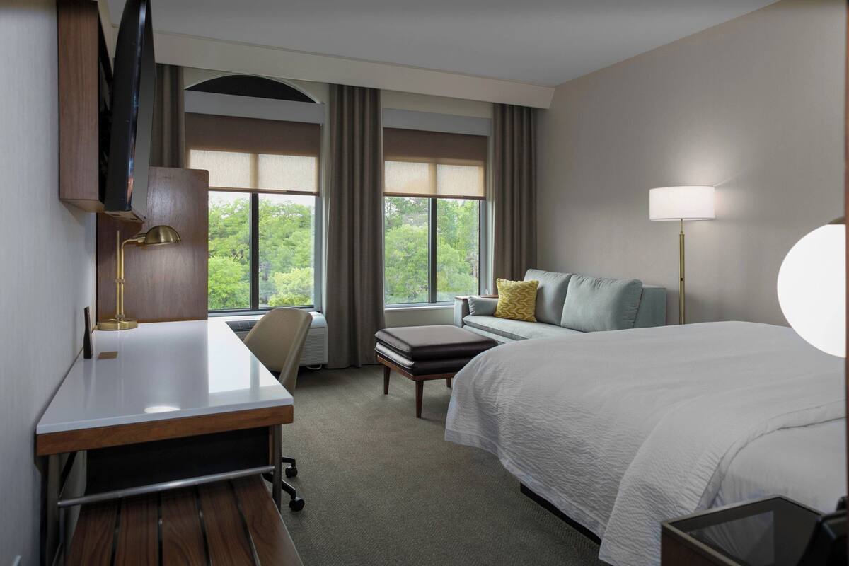 Habitación de hotel con cama, escritorio, silla y un ventanal con cortinas.