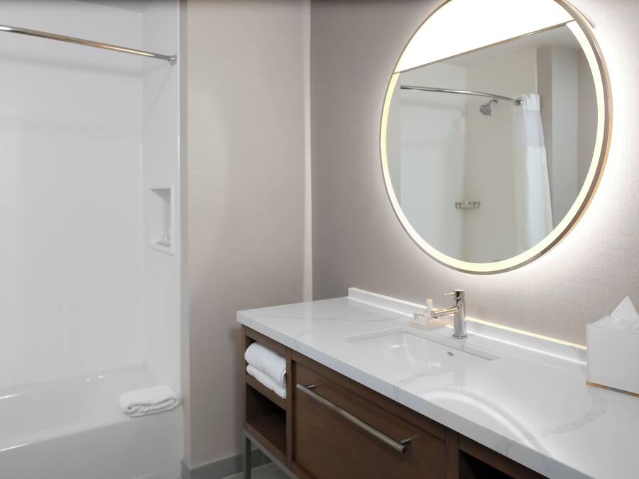 Une salle de bain moderne avec un lavabo blanc, un miroir rond doté d’un éclairage et une douche à parois de verre.