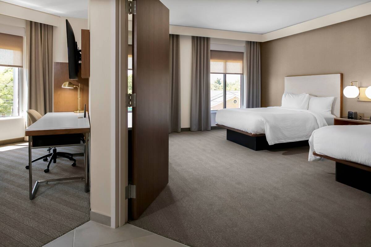 Habitación de hotel con escritorio, dos camas, televisor y vistas al exterior.