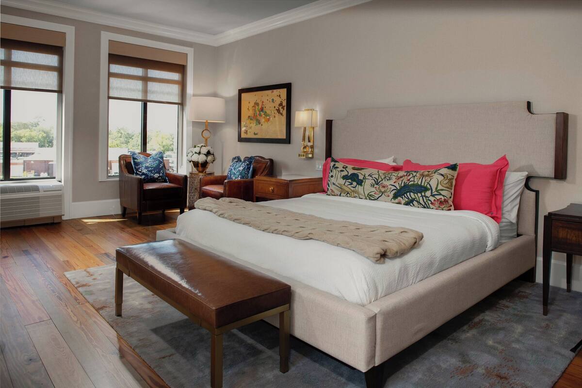 Un dormitorio amplio con una cama grande, un banco, un escritorio y una silla. Hay dos ventanas con persianas, una lámpara de pared y un cuadro enmarcado.
