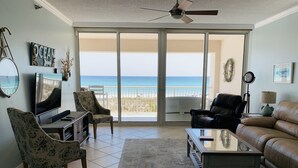 A living room with a TV, two chairs, a sofa, and a coffee table. There is a large window with a view of the beach.