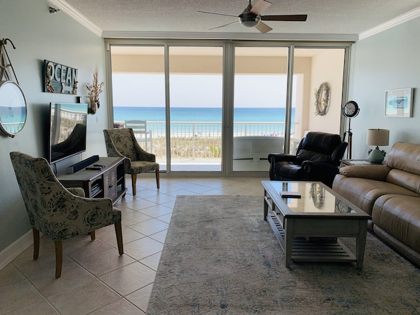 A living room with a TV, two chairs, a sofa, and a coffee table. There is a large window with a view of the beach.