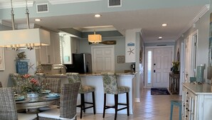 A spacious kitchen with a central island, a dining area with wicker chairs, and a hallway leading to a door.